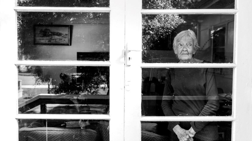 Black and white photo of elderly woman inside a house looking outside through a glass door.