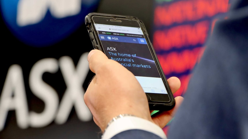一名男子手里拿着手机，背景是澳洲股市的牌价电子屏