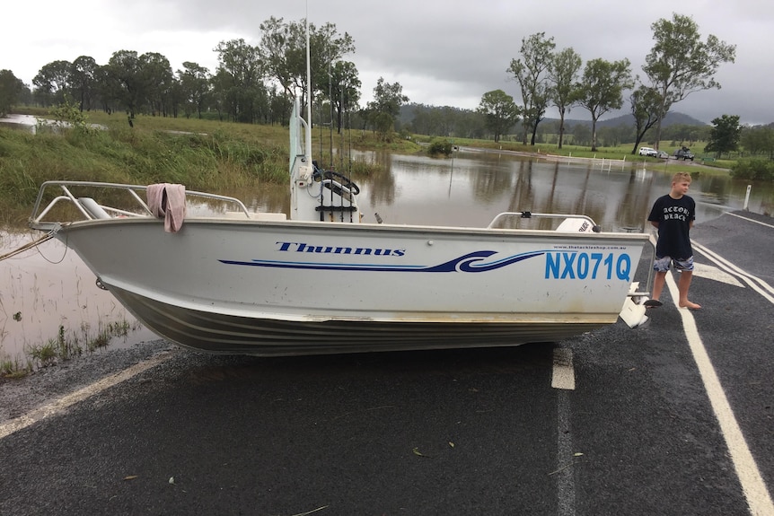 An image of a boat sitting on a bitumen road with flood water behind it