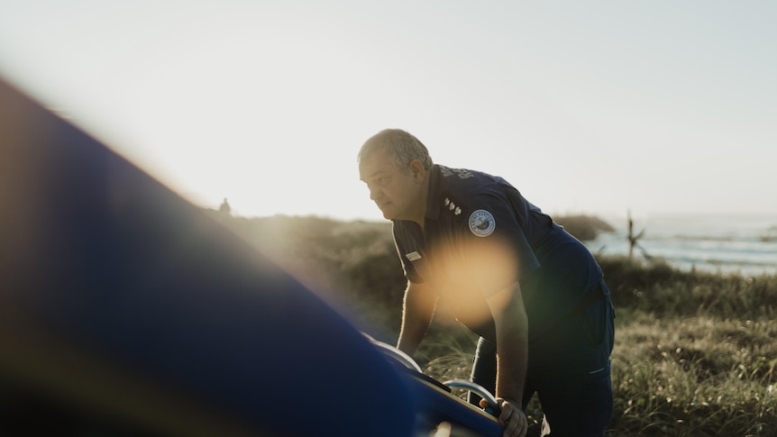 A man in blue uniform pushes the back end of a boat. There is shrubbery surrounding him.