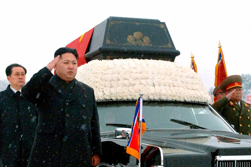 Kim Jong Un saltues while standing next to a hearse with a coffin on the roof. 