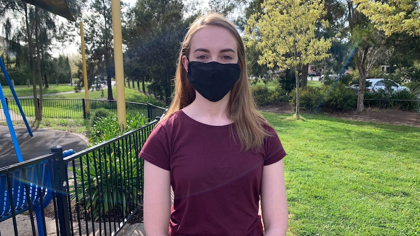 A woman wearing a red top and face mask looks at the camera 