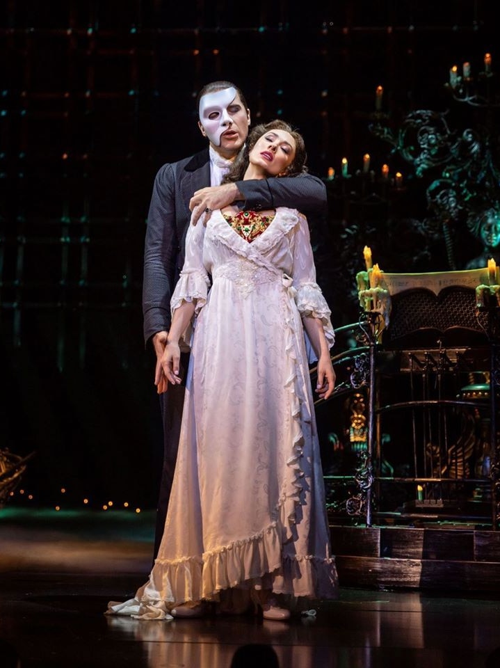 Claire Lyon stands on stage in Phantom of the Opera with the Phantom behind her.