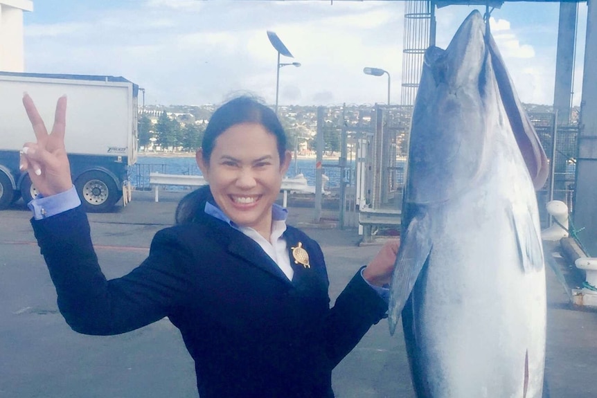 Woman on left holding up two fingers posing next to tuna being winched in air next to her