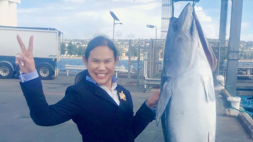 Woman on left holding up two fingers (the V sign) posing next to a tuna being winched into the air next to her.