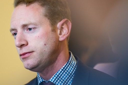 A man in navy suit and checked shirt looks off camera, with a sombre expression.