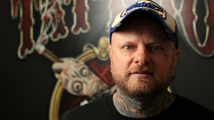 Lance Daly stares at the camera wearing a baseball cap, with visible tattoos on his neck and face.