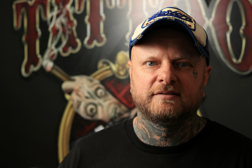 Lance Daly stares at the camera wearing a baseball cap, with visible tattoos on his neck and face.