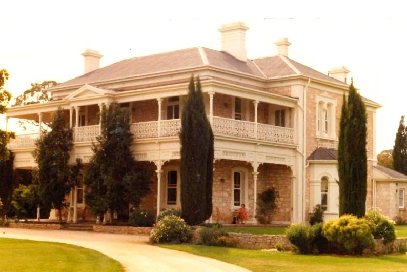 A beautiful two-storey old stone building, bordered by well kept trees and lawn.