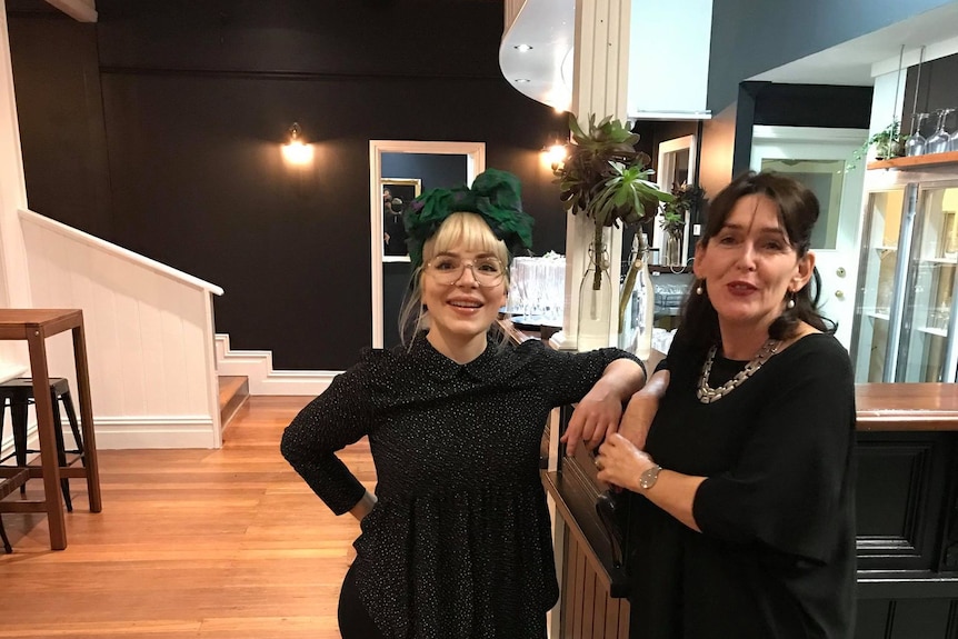 Two women stand at a bar in a restaurant, one with an oversized bow in her hair.