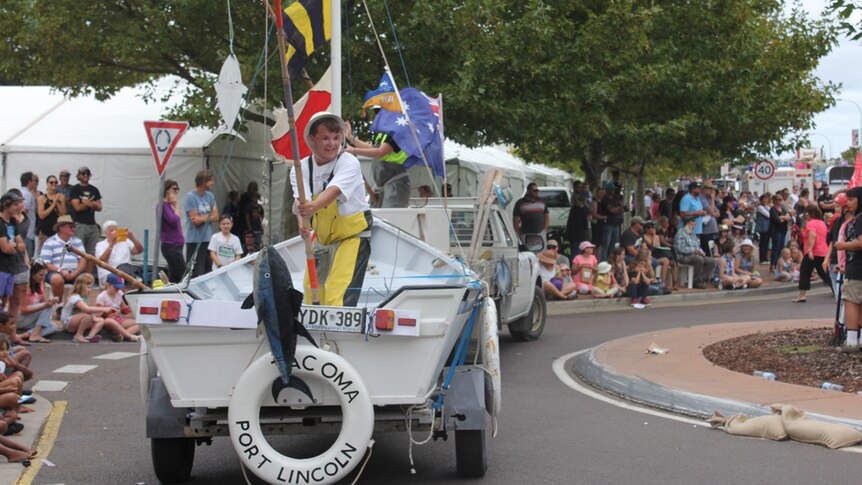 A man with a fishing rod holding a fake fish and standing on the back of a trailer parades through the streets of Port Lincoln.