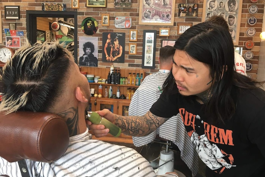 A hairdresser shaves a man's beard in a barber shop that has pictures hanging on the brick wall behind to two men.