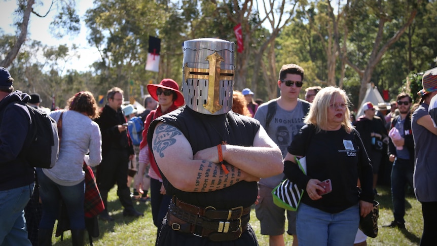 A man wearing a full-face metal helmet stands with this arms crossed amongst a crowd.