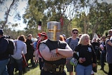 A man wearing a full-face metal helmet stands with this arms crossed amongst a crowd.