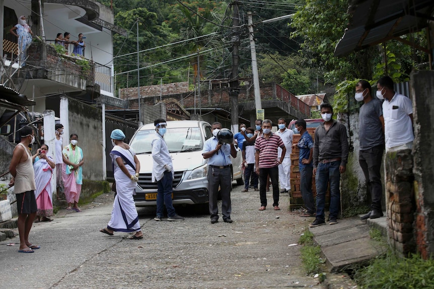 People wearing face masks watch health workers collect a patient and take them away in a van.