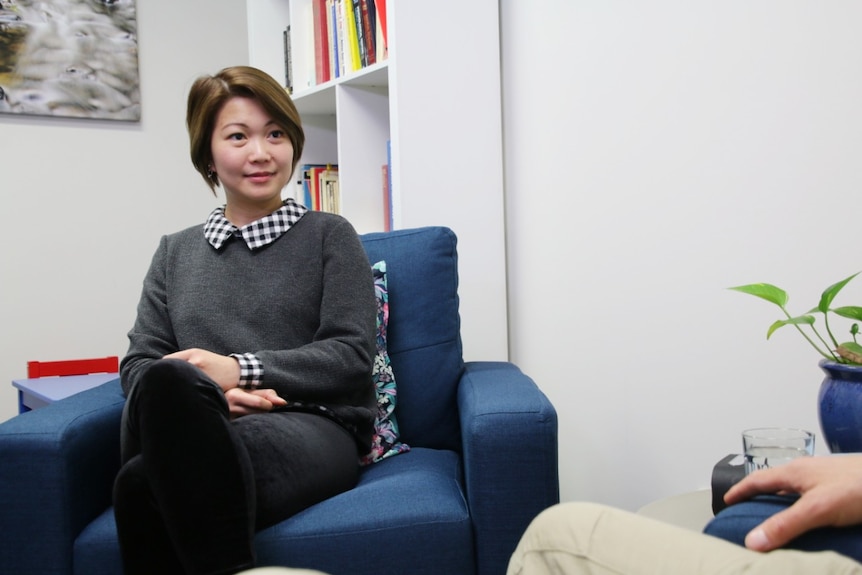 A woman with short brown hair wearing a jumper and a collared shirt sits on a blue armchair.