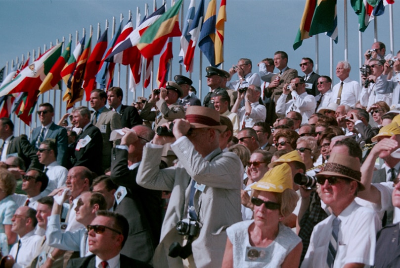 Crowd of spectators sitting and standing, some holding cameras on cloudless sunny day, with a row of flags on poles behind them.