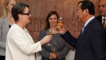 An Australian woman in white clinks glasses with a Cambodian man in a dark suit. A woman looks very uncomfortable in background.