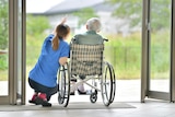 A generic image of a carer crouching down and pointing next to a man who is using a wheelchair.