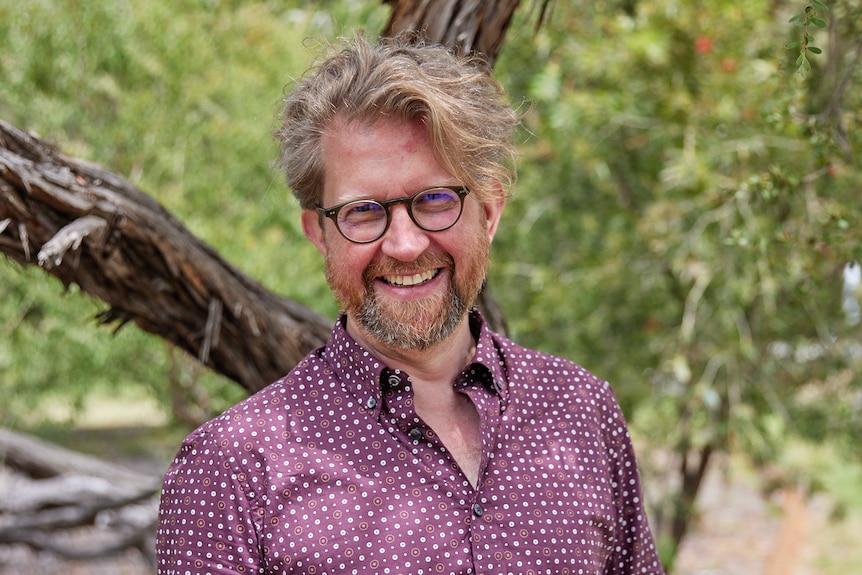A middle-aged man with light brown hair and glasses smiles. He is standing by a tree, wearing slacks and a smart shirt.