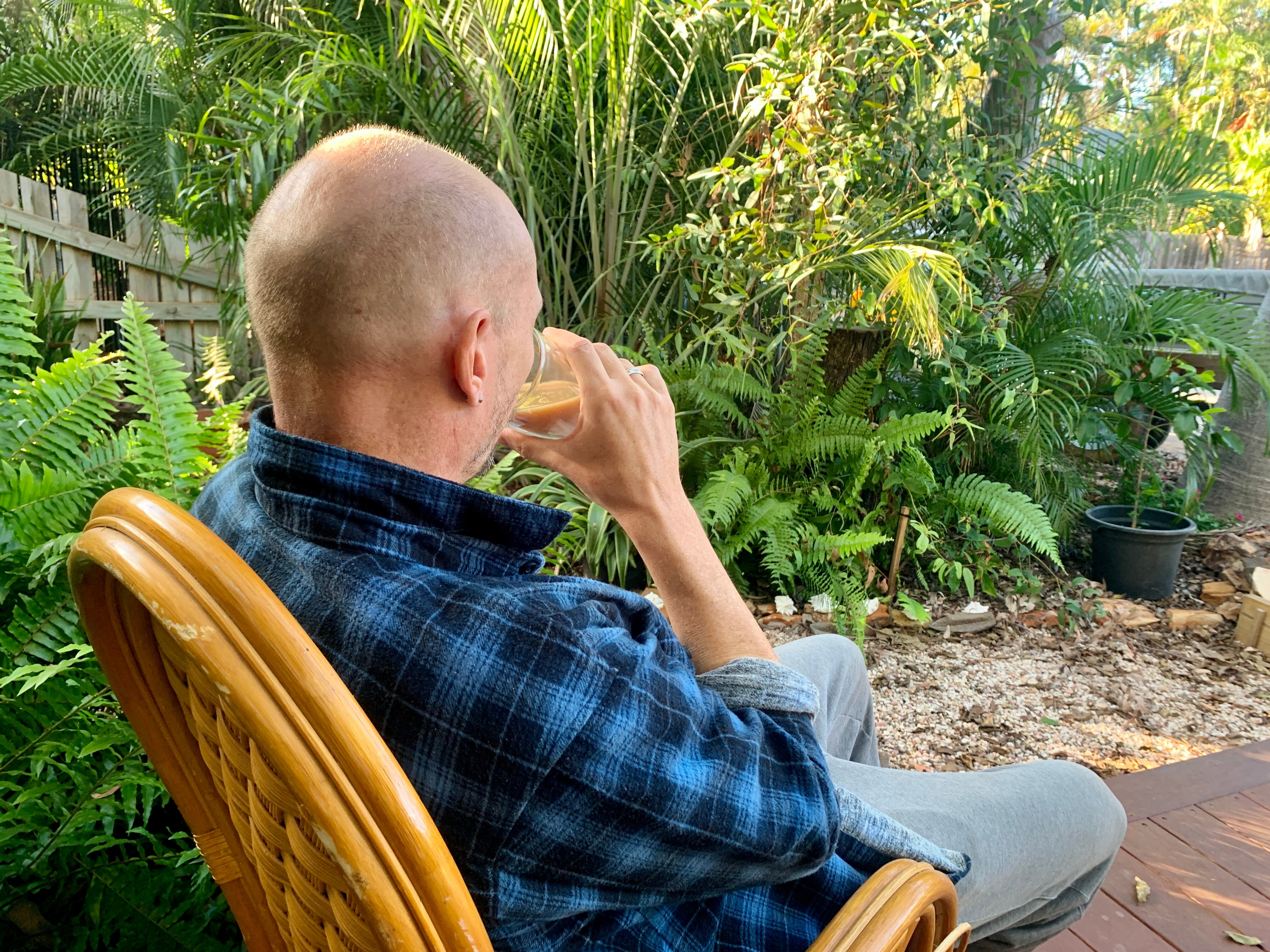 A bald man sitting alone in a cane chair in his back yard drinking a cup of tea.