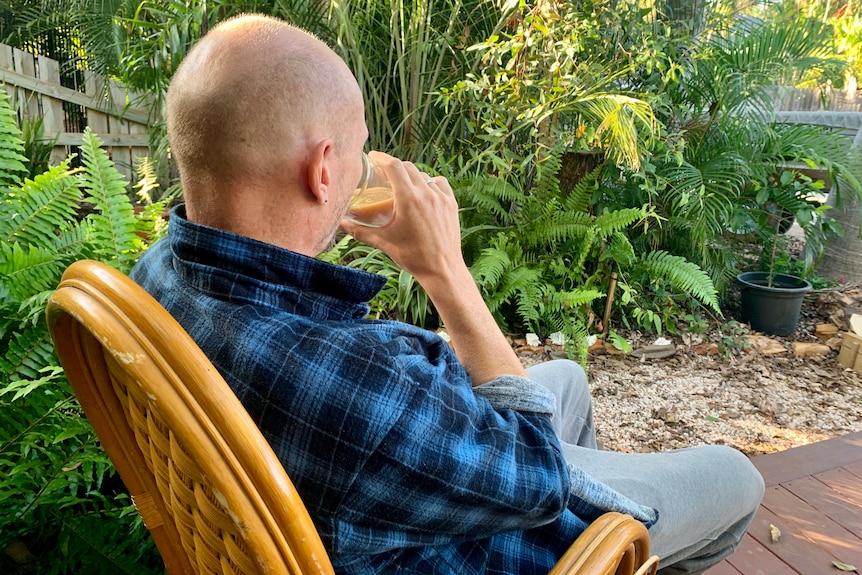 A bald man sitting alone in a cane chair in his back yard drinking a cup of tea.