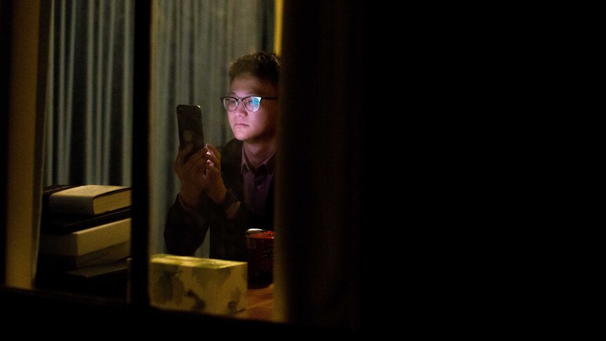 Johnny Zhang through a window, as he looks at his phone in his studio apartment