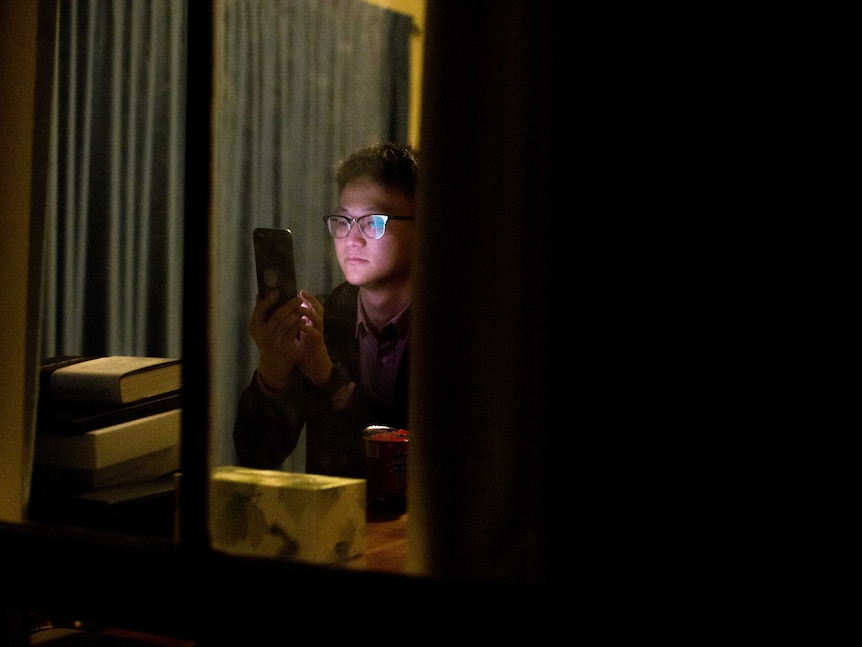 Johnny Zhang through a window, as he looks at his phone in his studio apartment