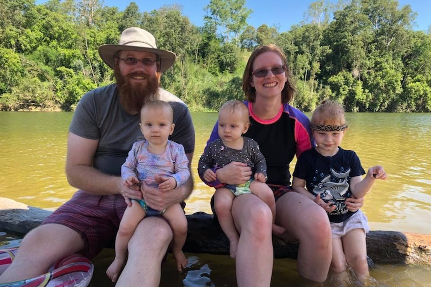 A mother, father and their three girls sit smiling in front of a creek.