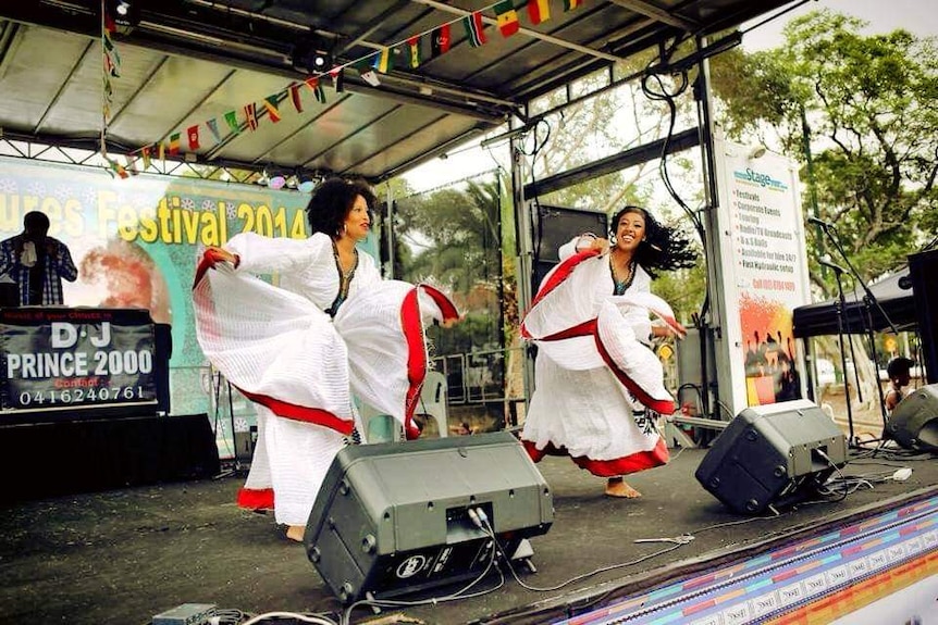 Two Ethiopian women dressed in flowing white dresses with red and green trim dancing on stage with a DJ behind them