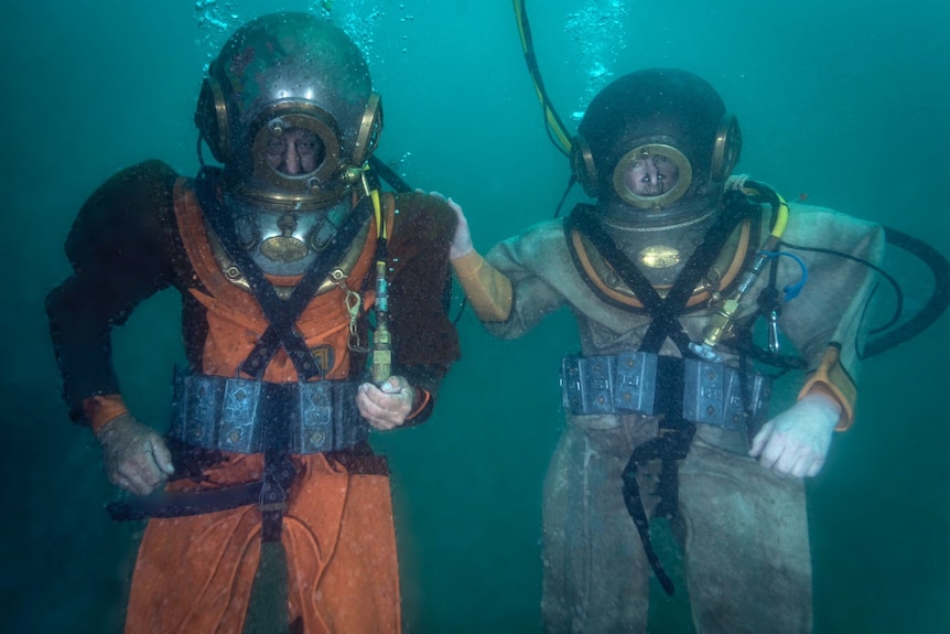 An underwater photo of two divers wearing weighted belts, helmets and canvas suits with oxygen bubbles drifting up. 