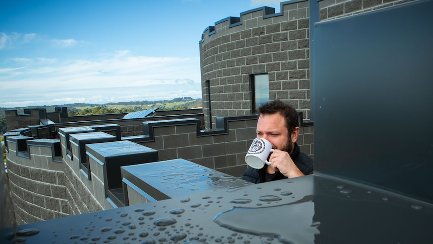A man sips from a coffee mug on a castle-like battlement, with grey, crenellated walls