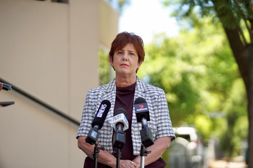 A woman wearing a purple and white blazer and purple shirt stands in front of news microphones