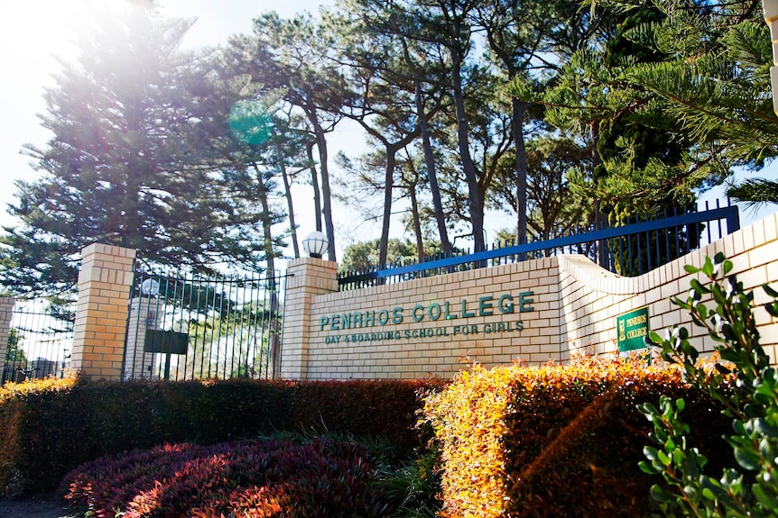 A sign saying 'Penrhos College' on a wall, with a fence on top and trees in the background'