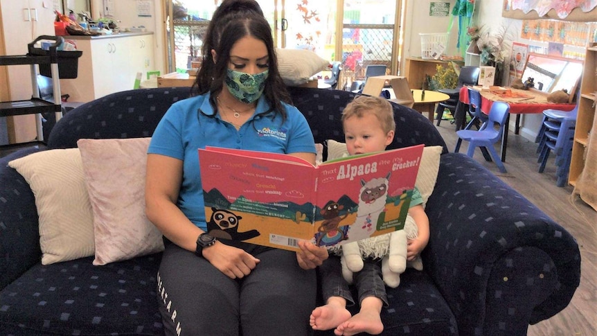 Educator, wearing a mask, and a child sitting on a couch reading a book