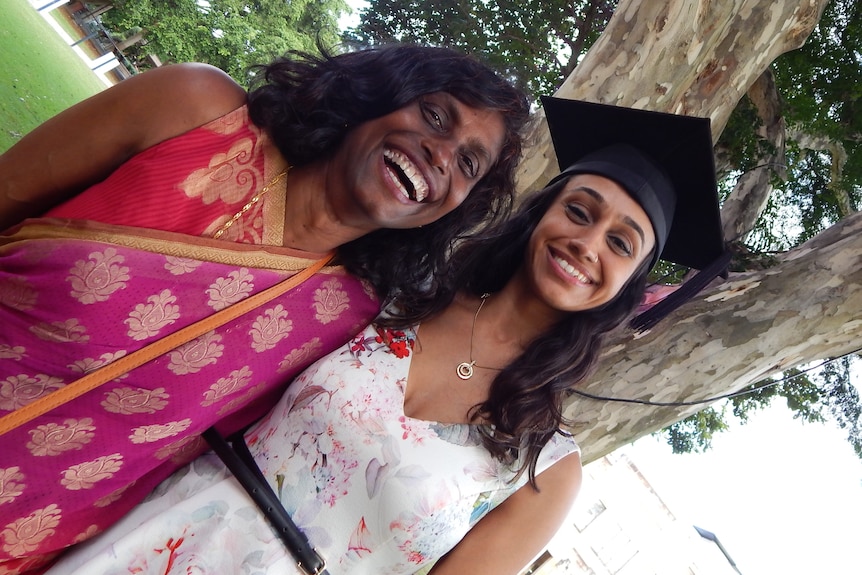 Indrani wearing a saree stands next to Tasha who is wearing a graduation cap, both smile looking at the camera.