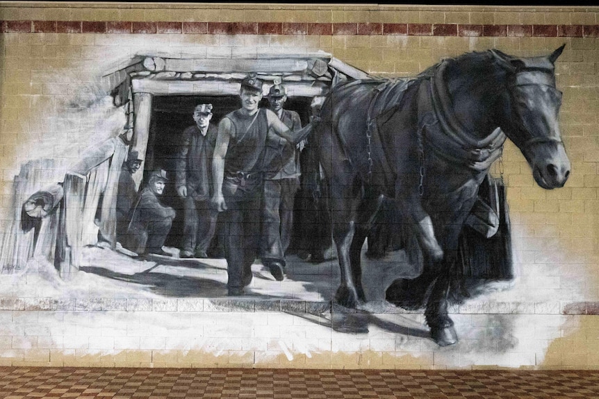 A black and white painting on a wall showing a horse next to a man in front of three other people in front of mine shaft.