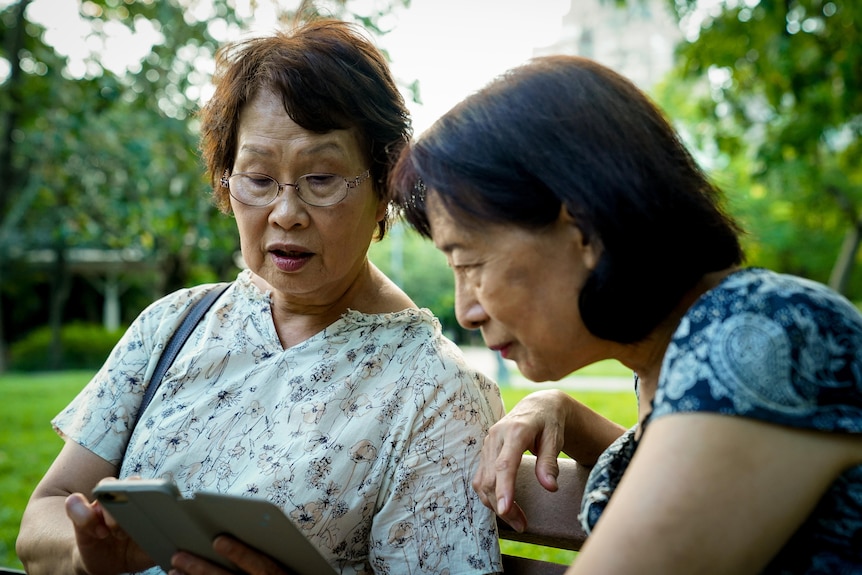 Two older women sit on a park bench, one showing the other her phone screen