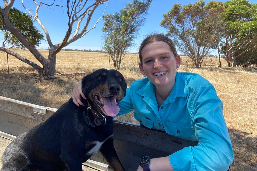 A young woman in a blue button up shirt smiles on a farm with her dog.