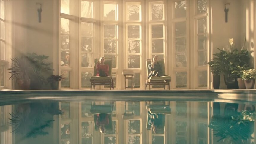 A woman in red and a woman in deep green sit by an indoor pool, smoking.