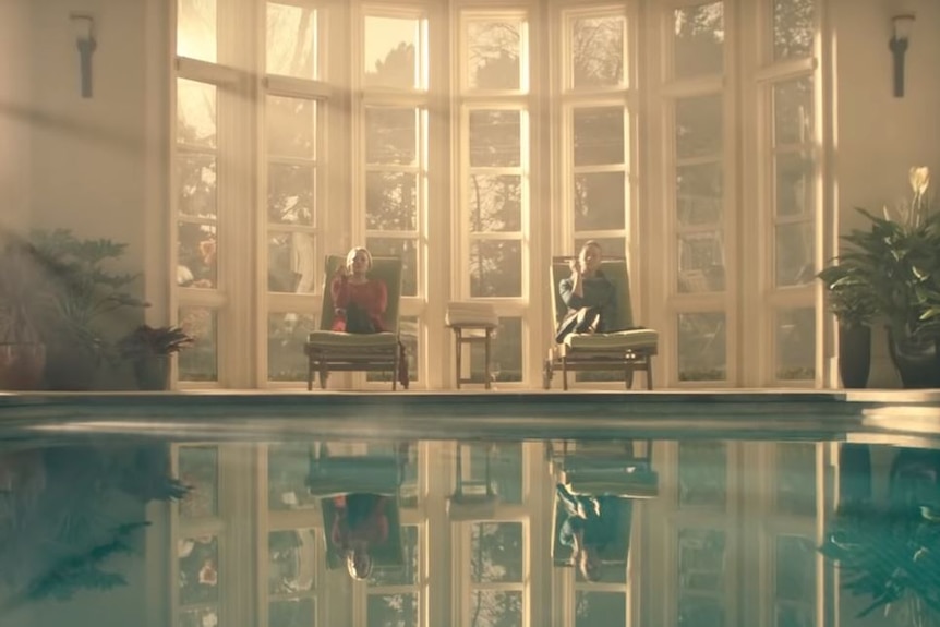 A woman in red and a woman in deep green sit by an indoor pool, smoking.