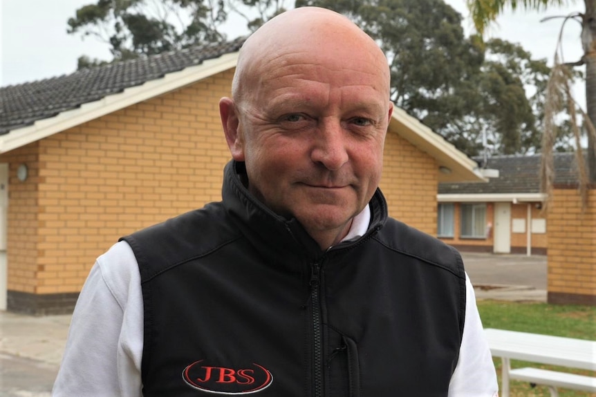 A middle-aged man wearing a black woollen vest with JBS embroidered is smiling in front of several yellow brick buildings