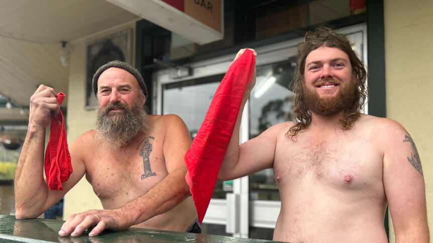 Two men without a shirt on hold up red handkerchiefs out the front of a pub