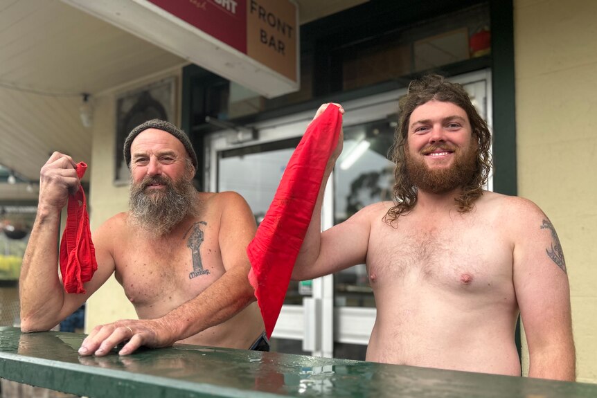 Two men without a shirt on hold up red handkerchiefs out the front of a pub
