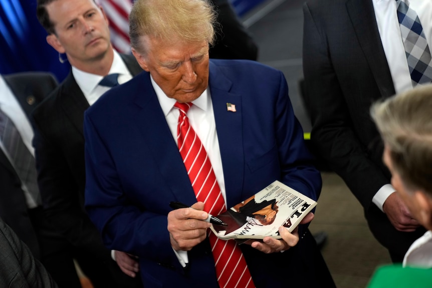 Donald Trump in a blue suit and tie signs a Playboy magazine cover at a rally in Iowa
