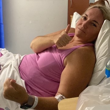 A woman in a pink tank top lying on a hospital bed giving a thumbs up