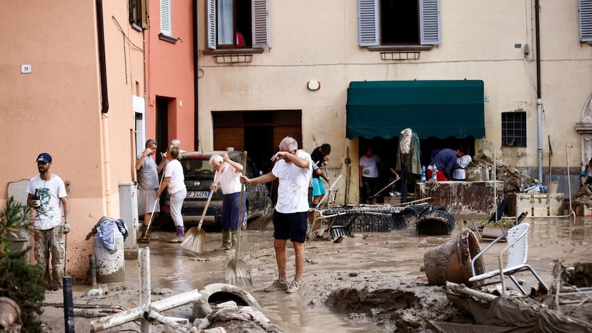 Man wipes his forehead as he holds a spade and is surrounded by mud and people cleaning it up. 
