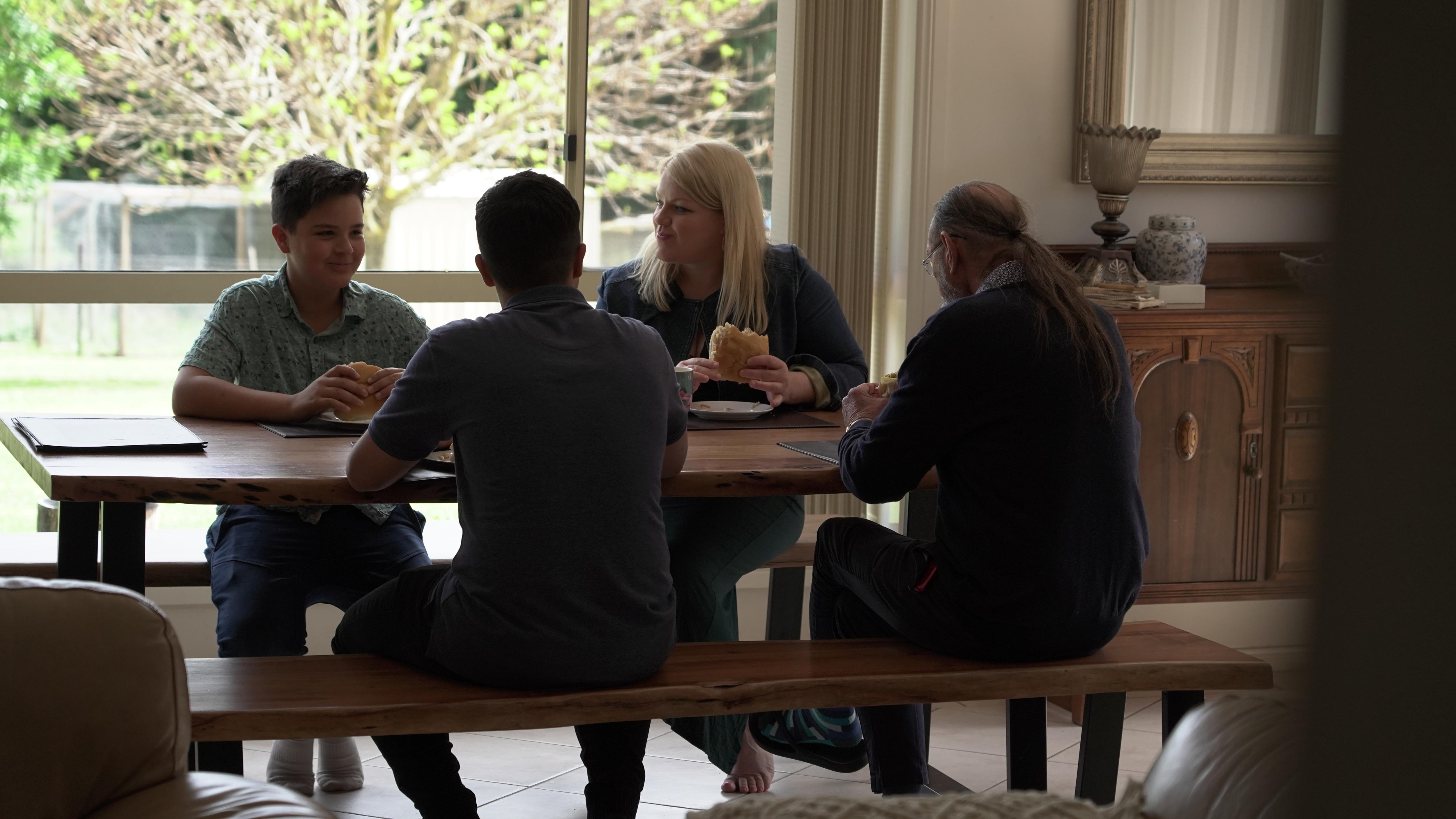 Catherine, Gregory, Charlton and Jackson sitting around a table eating burgers, large glass window behind them.