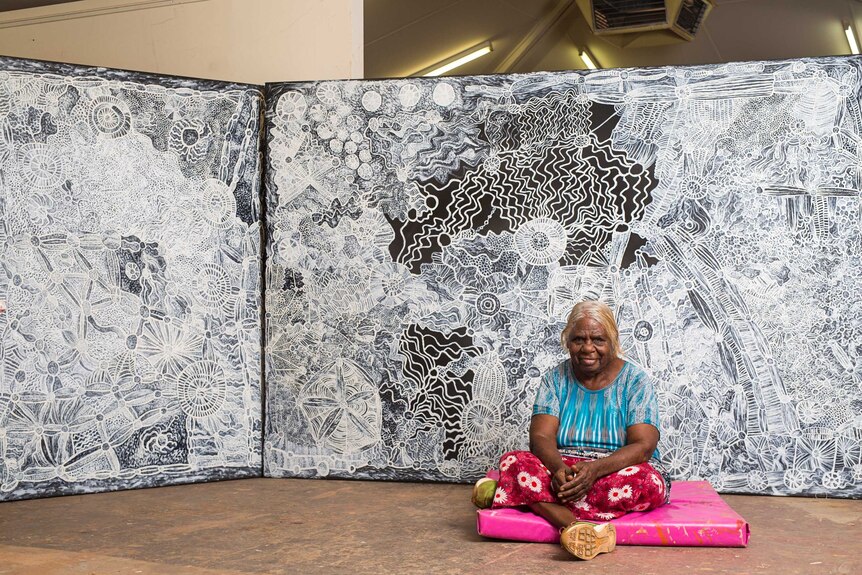 An older Aboriginal woman in colourful clothes sits in front of a vast detailed canvas painting in black and white