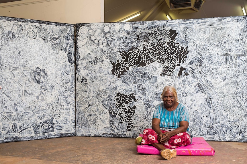 An older Aboriginal woman in colourful clothes sits in front of a vast detailed canvas painting in black and white